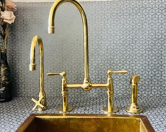 Op de afbeelding: Een gouden keukenkraan met twee handvatten en een aparte sproeikop. De kraan is gemonteerd op een aanrechtblad met een geometrisch patroon.