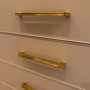 May include: Three gold drawer pulls with a rounded rectangular shape are mounted on a light pink dresser.