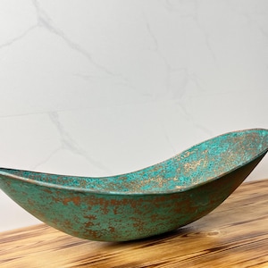 Könnte beinhalten: Eine handgefertigte, bootsförmige Dekoschale mit einem verwitterten türkis-bronzenen Finish. Die Schale ist aus Metall gefertigt und steht auf einer Holzoberfläche. Die Schale ist ca. 50 cm lang und 20 cm breit.