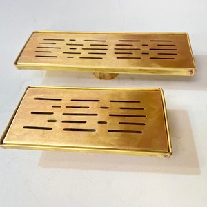 May include: Two gold-colored metal drain covers with rectangular shapes and a grid pattern of small holes. The drain covers are made of a shiny metal and have a slightly raised edge.