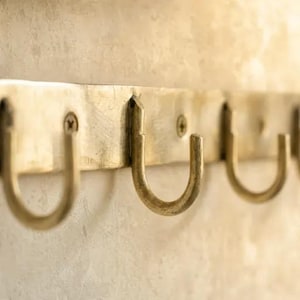 Puede incluir: Un perchero de pared horizontal de latón con múltiples ganchos en forma de U unidos a una barra de metal plana. El latón tiene un acabado metálico cepillado con cabezas de tornillo visibles que aseguran la barra a la pared.