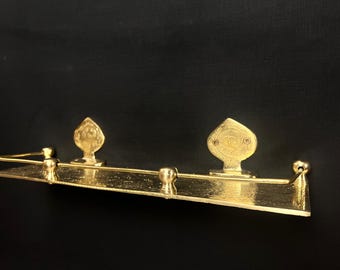 May include: A gold-colored, decorative metal shelf with two ornate, leaf-shaped accents and a central, rounded knob. The shelf is rectangular with a slightly angled design, and the overall aesthetic is vintage.