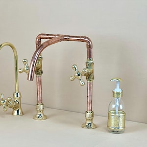 以下が含まれることがあります： 2つのハンドルと別々のウォーターフィルター蛇口が付いた銅と真鍮製のキッチン蛇口。蛇口は高くアーチ型の注ぎ口があり、カウンタートップに取り付けられています。画像には、金の石鹸ディスペンサーも写っています。