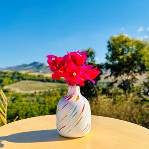 May include: A white ceramic vase with a rounded body and a narrow neck, decorated with diagonal brushstrokes in blue, yellow, orange, and red. The vase holds a vibrant bouquet of bright pink bougainvillea flowers with small white centres.