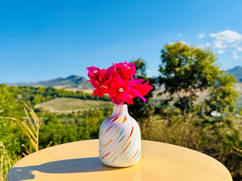May include: A white ceramic vase with a rounded body and a narrow neck, decorated with diagonal brushstrokes in blue, yellow, orange, and red. The vase holds a vibrant bouquet of bright pink bougainvillea flowers with small white centres.