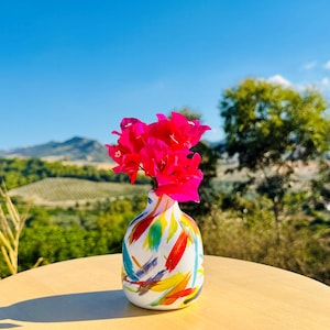 May include: A small, rounded ceramic vase with a white base featuring colourful, abstract brushstrokes in red, blue, yellow, and green. The vase holds a cluster of vibrant, bright pink bougainvillea flowers extending from the narrow neck opening.