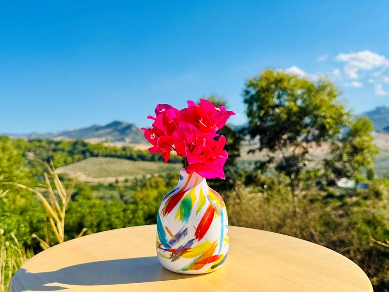 May include: A small, rounded ceramic vase with a white base featuring colourful, abstract brushstrokes in red, blue, yellow, and green. The vase holds a cluster of vibrant, bright pink bougainvillea flowers extending from the narrow neck opening.