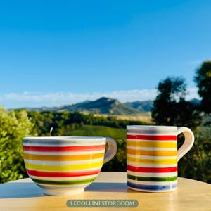 Könnte beinhalten: Zwei Keramikbecher mit horizontalen Streifen in Rot, Gelb, Grün und Blau. Einer ist ein großer schalenförmiger Becher, der andere ein Standardbecher. Die Becher stehen auf einer Holzoberfläche mit einem malerischen Hintergrund aus Bergen und blauem Himmel.