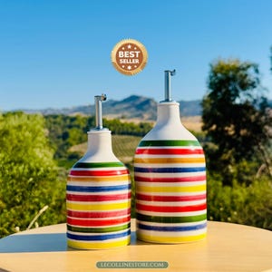 May include: Two ceramic oil dispensers with a white base and horizontal stripes in red, yellow, green, and blue. Each bottle features a silver-coloured metal pour spout. A gold-coloured circular badge with the text BEST SELLER is visible above.