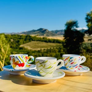May include: Three white ceramic espresso cups and saucers with colourful abstract designs. One cup has red, yellow, and blue paint strokes, another has green brushstrokes, and the third has colourful feather-like shapes.