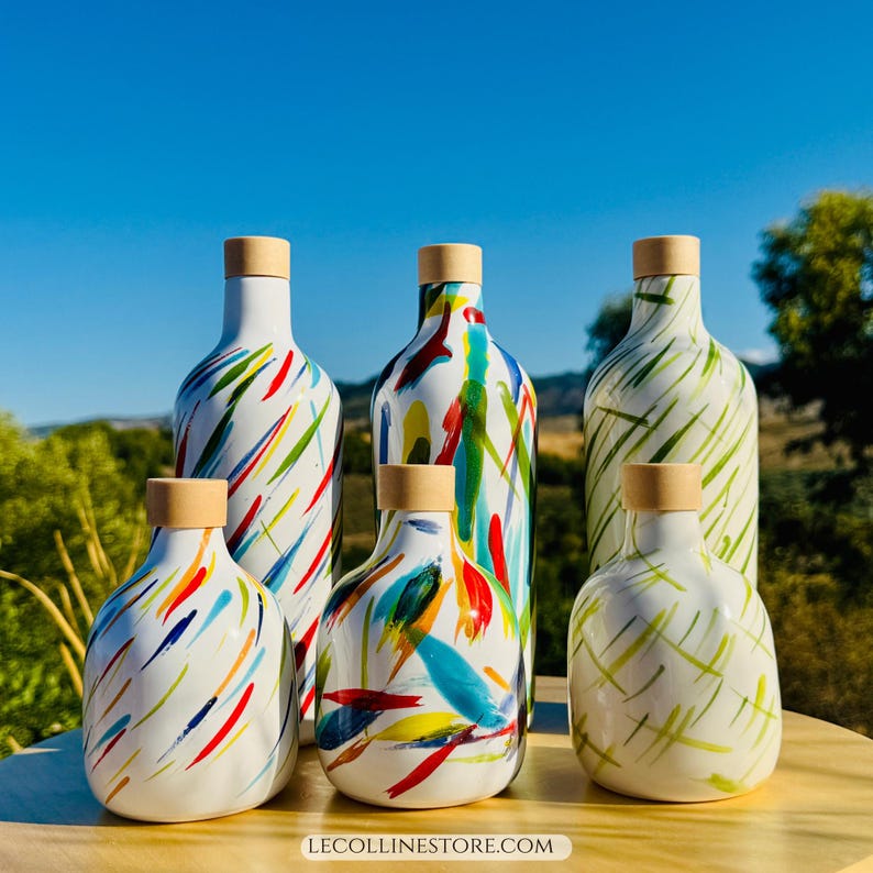 Sei bottiglie in ceramica bianca con tappi in legno. Sono decorate con pennellate astratte colorate in rosso, blu, giallo e verde. Le bottiglie variano per dimensioni e forma e sono disposte su una superficie di legno.