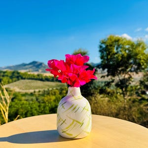 May include: A small, rounded white ceramic vase featuring a pattern of thin, light green brushstrokes. The vase contains a cluster of vibrant, bright pink bougainvillea flowers, which extend upwards from the narrow opening at the top.