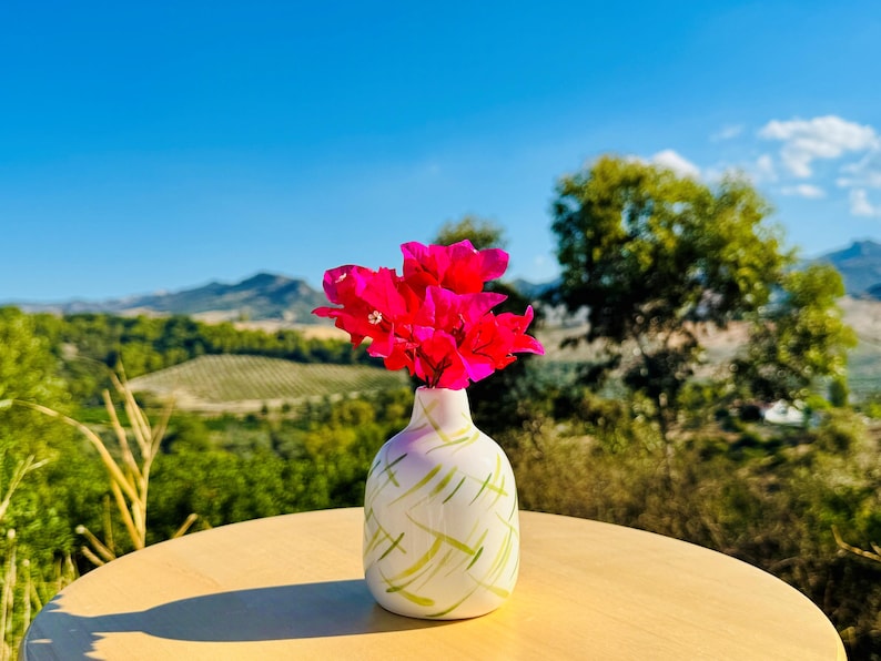 May include: A small, rounded white ceramic vase featuring a pattern of thin, light green brushstrokes. The vase contains a cluster of vibrant, bright pink bougainvillea flowers, which extend upwards from the narrow opening at the top.