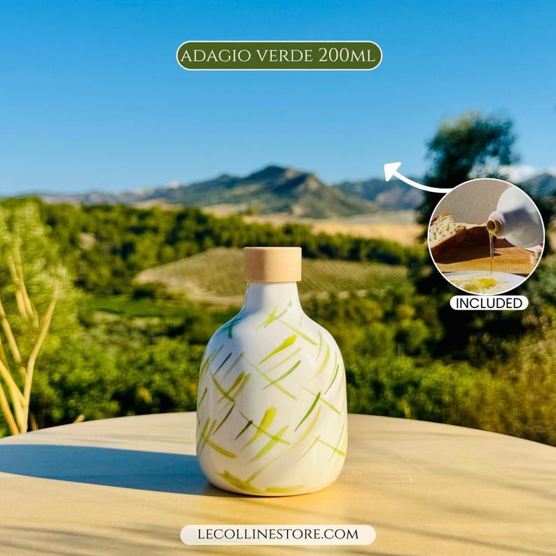 May include: A white ceramic bottle with a light green abstract brushstroke pattern and a cylindrical wooden stopper. The text ADAGIO VERDE 200ML is visible at the top, and a circular inset shows the bottle pouring oil onto bread.