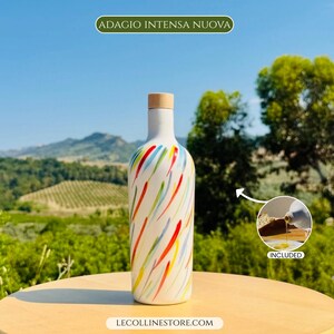 May include: A white bottle with a light wood-coloured cap, featuring a colourful pattern of red, blue, and green brushstrokes. The text ADAGIO INTENSA NUOVA appears at the top, and LECOLLINESTORE.COM is at the bottom. An inset shows oil pouring.