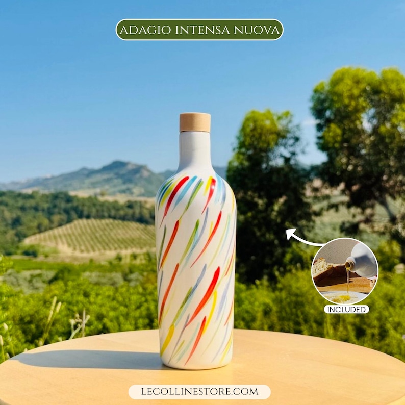 May include: A white bottle with a light wood-coloured cap, featuring a colourful pattern of red, blue, and green brushstrokes. The text ADAGIO INTENSA NUOVA appears at the top, and LECOLLINESTORE.COM is at the bottom. An inset shows oil pouring.