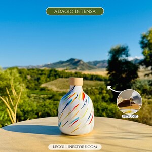 May include: A white ceramic bottle with a rounded shape and a light wood-coloured stopper, featuring a colourful pattern of diagonal brushstrokes in red, blue, yellow, and green. The text ADAGIO INTENSA and LECOLLINESTORE.COM are visible.