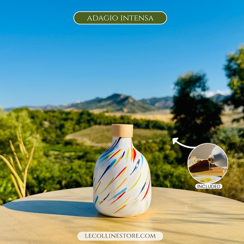 May include: A white ceramic bottle with a rounded shape and a light wood-coloured stopper, featuring a colourful pattern of diagonal brushstrokes in red, blue, yellow, and green. The text ADAGIO INTENSA and LECOLLINESTORE.COM are visible.