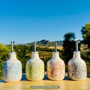 以下が含まれることがあります： ステンレス製の注ぎ口が付いた、4つのセラミックオイルボトル。各ボトルは白で、青、緑、赤、黒のユニークな斑点模様があります。ボトルは、山と青い空の風光明媚な背景を持つ木製の表面に配置されています。