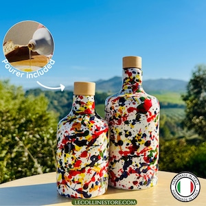 Può includere: Due bottiglie decorative in vetro con tappi in legno. Le bottiglie sono bianche con schizzi di vernice colorati in rosso, giallo, blu e nero. L'immagine include un piccolo inserto che mostra un versatore. Il testo "LECOLLINESTORE.COM" è in basso.