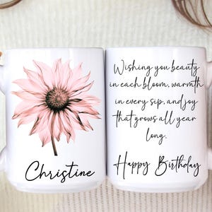 May include: Two white ceramic mugs. One mug features a pink flower and the name "Christine". The other mug has the text "Wishing you beauty in each bloom, warmth in every sip, and joy that grows all year long. Happy Birthday."