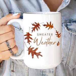 May include: A white ceramic mug featuring a circular arrangement of brown autumn oak leaves. The text "SWEATER Weather" is printed in the center in a combination of dark brown block letters and a light brown script font.