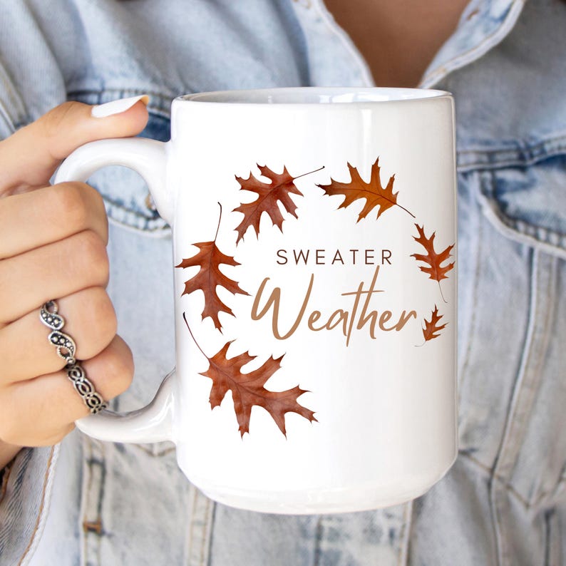 May include: A white ceramic mug featuring a circular arrangement of brown autumn oak leaves. The text "SWEATER Weather" is printed in the center in a combination of dark brown block letters and a light brown script font.