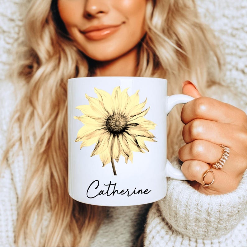 May include: White ceramic mug with a yellow sunflower design and the name "Catherine" in cursive. The sunflower has a dark centre. The mug has a white handle. The design is printed on the front.