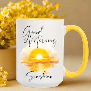 May include: White ceramic mug with a yellow handle and rim. The mug features the words "Good Morning Sunshine" in black script, along with a watercolor image of a sunrise over water. A cheerful and bright design.