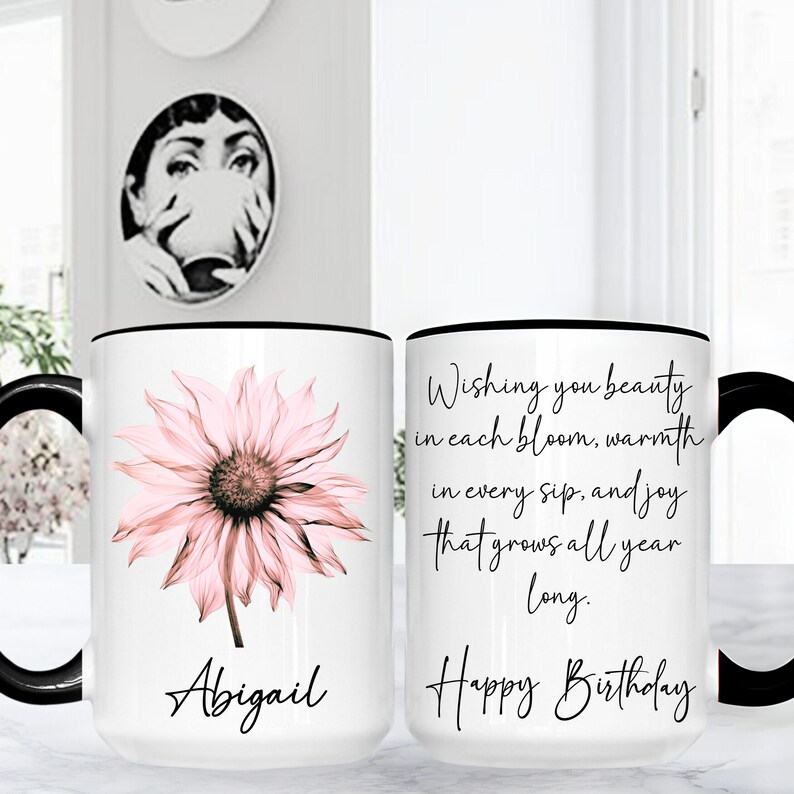 May include: White ceramic mug with a black handle and rim, featuring a pink flower design and the name "Abigail." The other side has the text "Wishing you beauty...Happy Birthday."