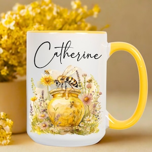 May include: White ceramic mug with a yellow handle and rim. The mug features a watercolor illustration of a honey pot with a bee, surrounded by flowers. The name "Catherine" is written in black script.