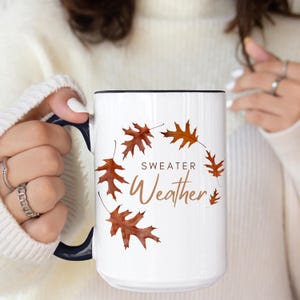 May include: A white ceramic mug with a dark blue handle and interior. The mug features a circular arrangement of brown oak leaves and the text "SWEATER Weather" printed in a combination of dark brown block letters and a brown script font.