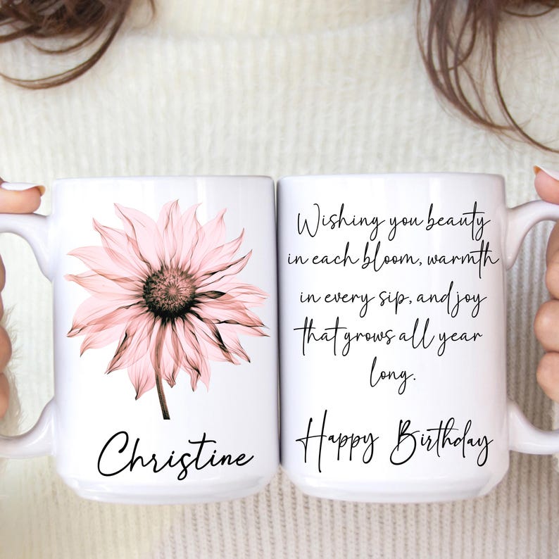 May include: Two white ceramic mugs. One mug features a pink flower and the name "Christine". The other mug has the text "Wishing you beauty in each bloom, warmth in every sip, and joy that grows all year long. Happy Birthday."