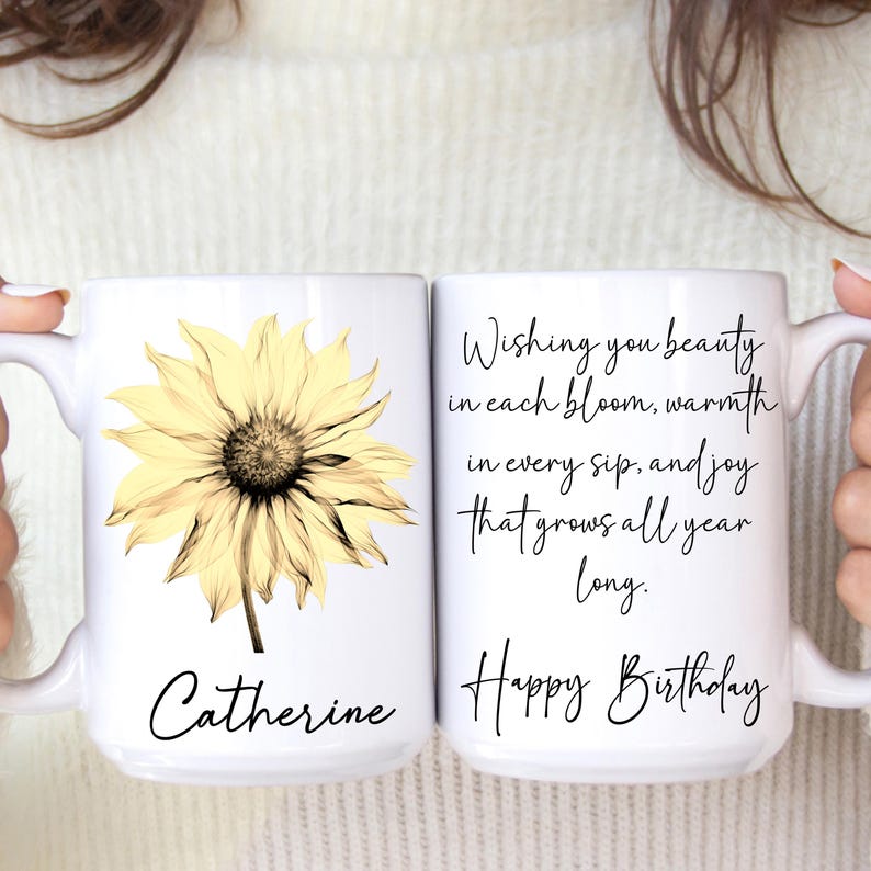 May include: Two white ceramic mugs. One mug features a yellow sunflower graphic and the name "Catherine". The other mug has the text "Wishing you beauty in each bloom, warmth in every sip, and joy that grows all year long. Happy Birthday."