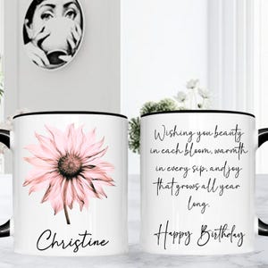 May include: White ceramic mug with a black handle and rim, featuring a pink flower design and the name "Christine." The other side has the text "Wishing you beauty...Happy Birthday."