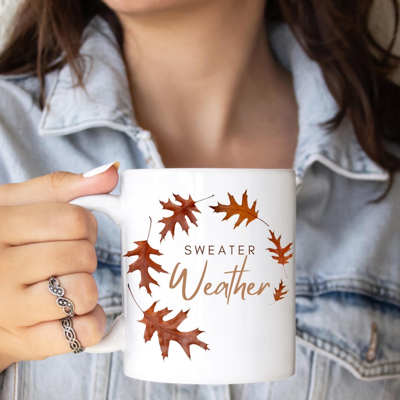 May include: A white ceramic mug featuring a circular arrangement of brown autumn oak leaves. The text "SWEATER Weather" is printed in the center in a combination of dark brown block letters and a light brown cursive script font.