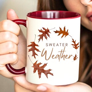 May include: A white ceramic mug with a deep maroon interior and handle. The exterior features a circular arrangement of brown oak leaves surrounding the brown text "SWEATER Weather" printed in a combination of block and script fonts.