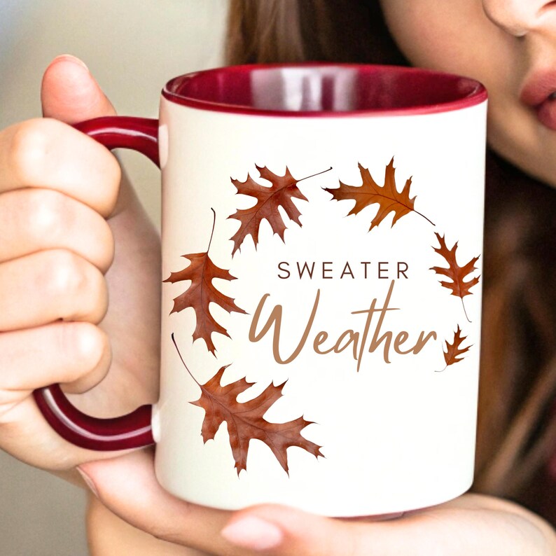 May include: A white ceramic mug with a deep maroon interior and handle. The exterior features a circular arrangement of brown oak leaves surrounding the brown text "SWEATER Weather" printed in a combination of block and script fonts.