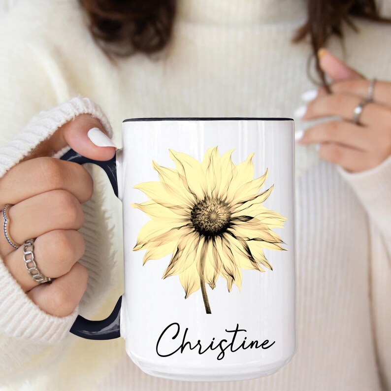 May include: White ceramic mug with a yellow sunflower design and a dark brown center. The mug has a black rim and handle, with the name "Christine" in a script font below the flower.