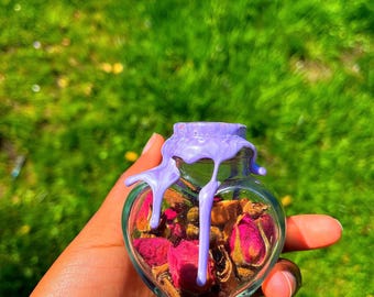 Peut inclure: Un bocal en verre en forme de cœur rempli de pétales de rose séchées et d'autres plantes. Le bocal est scellé avec un sceau de cire violet qui a coulé sur les côtés.