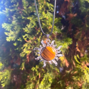 May include: A gold sun pendant necklace with a tiger's eye stone in the centre. The necklace is on a black cord and is hanging from a tree branch.