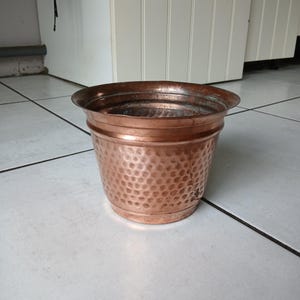 Może przedstawiać: Miedziana donica o młotkowanej fakturze. Donica ma szeroki, lekko rozszerzający się brzeg i cylindryczny korpus. Miedź ma ciepły, czerwonobrązowy odcień, a powierzchnia pokryta jest małymi, równomiernie rozmieszczonymi wgłębieniami.