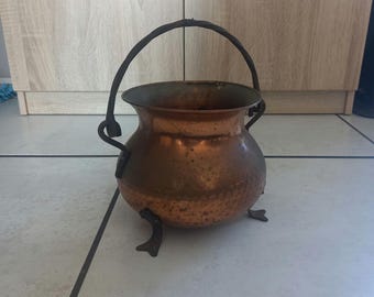 Puede incluir: Una olla de cobre con un acabado martillado y un asa de metal negro. La olla tiene tres patas y está sentada en un suelo de baldosas.