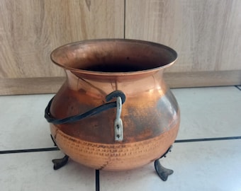 Puede incluir: Un caldero de cobre con un asa oscura y tres patas pequeñas. La olla tiene un cuerpo redondeado y una abertura ancha. El cobre tiene un color marrón rojizo cálido con algunas áreas más oscuras, lo que sugiere edad o uso. El caldero probablemente se usa para cocinar o con fines decorativos.