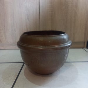 Op de afbeelding: Een gehamerde koperkleurige plantenbak met een afgeronde body en een brede, verhoogde rand. De plantenbak heeft een donker interieur en staat op een betegelde vloer. De koperen afwerking geeft het een rustieke, antieke uitstraling.