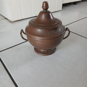 Puede incluir: Un recipiente de color cobre con tapa, dos asas laterales y una base redondeada. La tapa tiene un pequeño pomo redondeado. El recipiente está sobre una superficie embaldosada. El diseño general es vintage y decorativo.