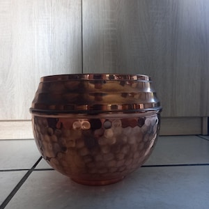 Pode incluir: Uma tigela de metal martelado cor de cobre com tampa. A tigela tem uma base arredondada e uma superfície texturizada. A tampa é de um tom mais escuro de cobre e tem um acabamento liso e polido. A tigela está sobre uma superfície de azulejos.