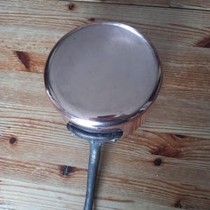 May include: A copper-colored saucepan with a long, dark metal handle. The pan has a rounded shape and a shiny surface. The handle is attached to the pan with rivets. The cookware is likely used for cooking on a stovetop.