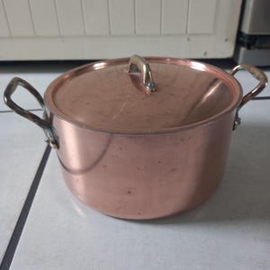 Puede incluir: Una olla de cobre con tapa y dos asas. La olla es brillante y tiene un asa de latón.