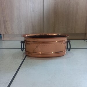 Pode incluir: Um vaso de plantas ovalado cor de cobre com duas alças pretas. O vaso tem um acabamento brilhante e uma borda ligeiramente elevada. Está sobre um chão ladrilhado.